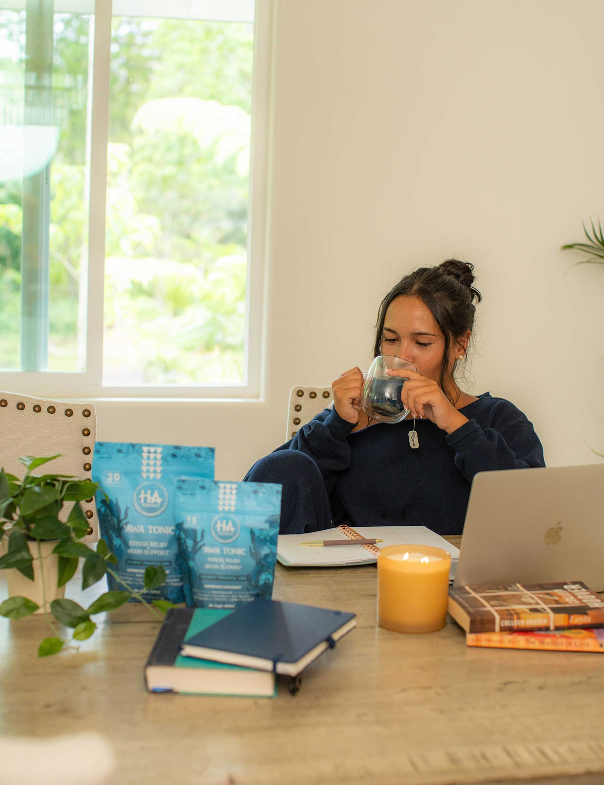 Woman sits at a table with a laptop, notebooks, a candle, and wellness drink packets, sipping from a glass mug. A plant and window are visible in the background.