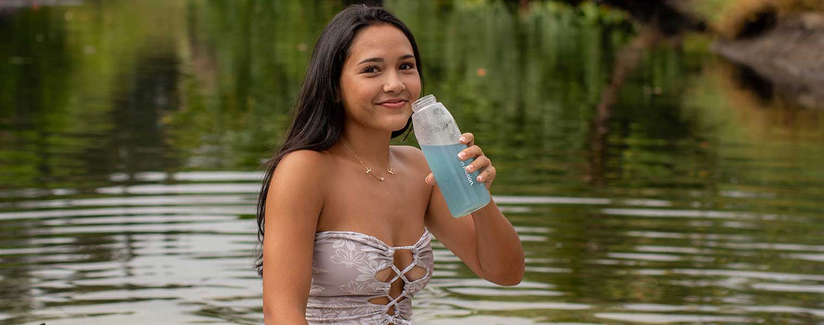 A woman in a patterned swimsuit sits by a body of water, holding and drinking from a glass bottle filled with blue spirulina tonic. Trees and greenery are in the background.
