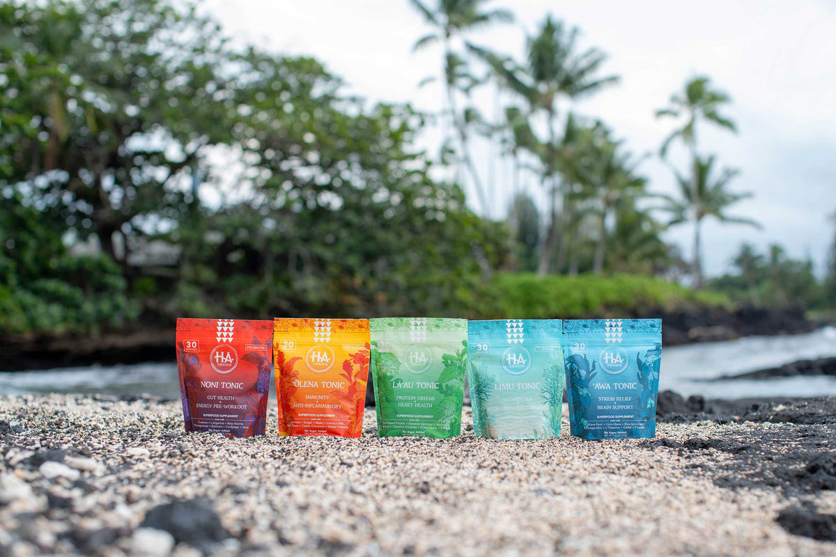 Five colorful HA Tonics pouches are lined up on a pebbled beach with palm trees and greenery in the background.