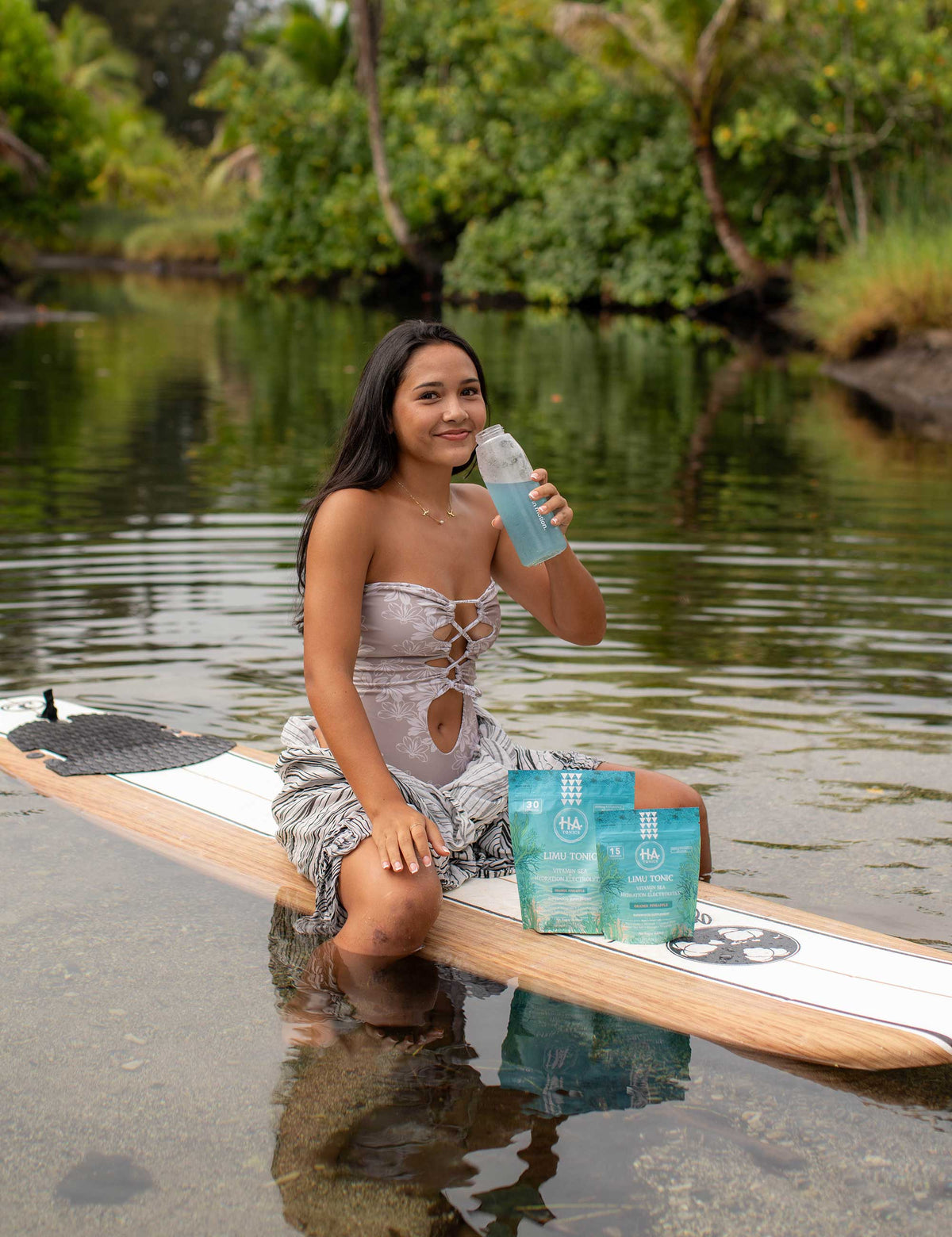 A woman kneels on a paddleboard in shallow water, holding a bottle with blue liquid and sitting beside a package labeled HA Tonics. Trees line the background.