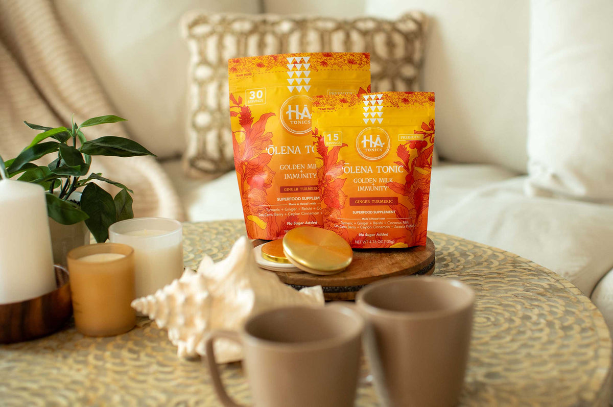 Two orange HA Tonics turmeric supplement bags on a table with candles, a seashell, a plant, two mugs, and a gold tin in a cozy living room setting.