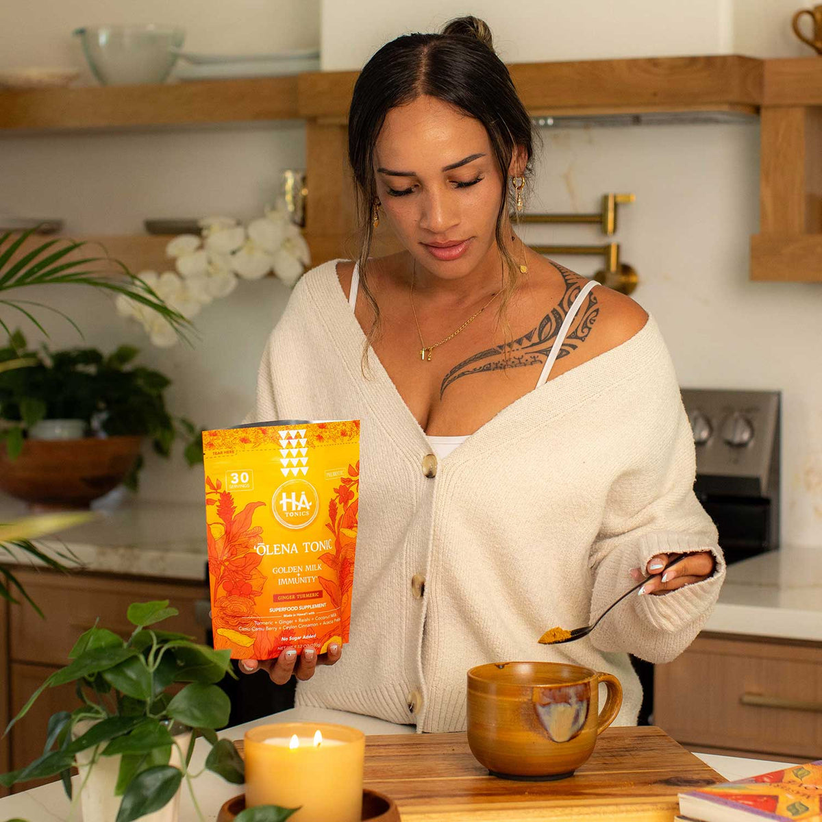 A woman stands in a kitchen holding a spoon and a packet labeled HA Olena Tonic, preparing a drink in a brown mug. A candle and plants are visible on the counter.