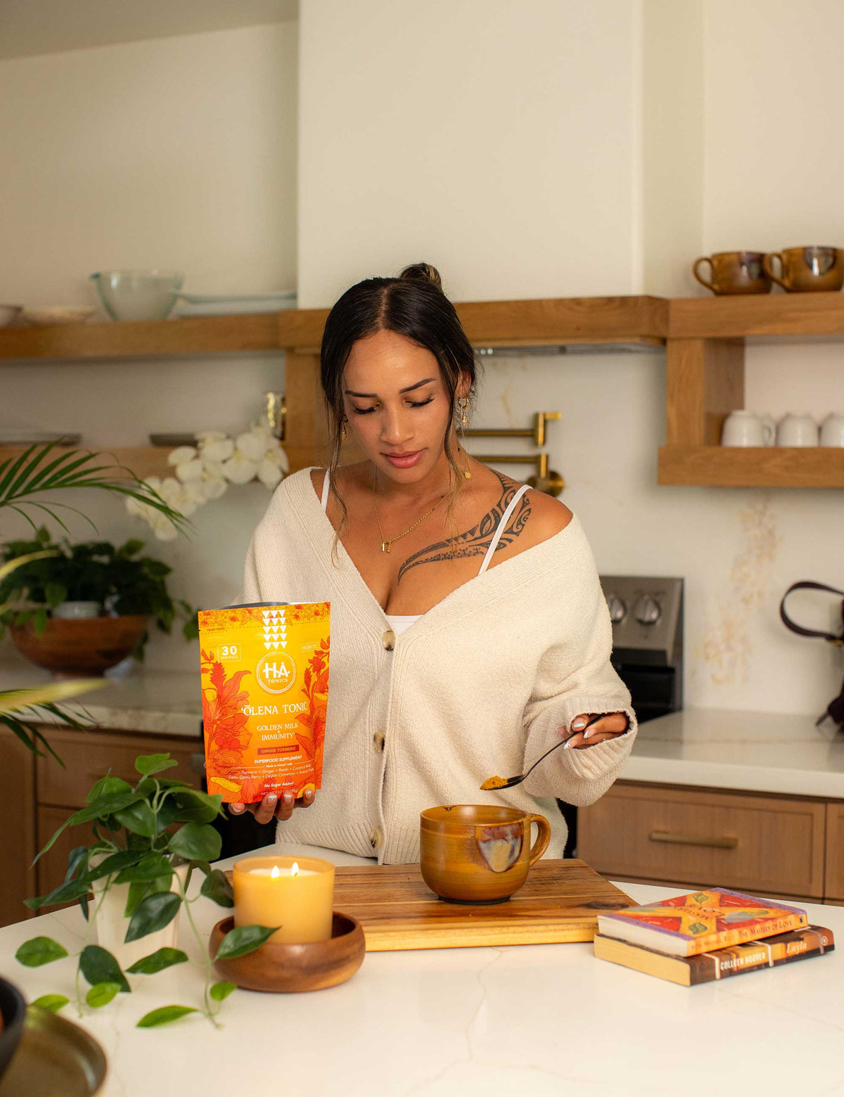 Woman in a kitchen scooping powder from an orange packet labeled Olena Tonic into a mug, with a candle, plant, and book on the counter.